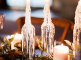 a wedding reception table with a greenery and pinecone table runner, pillar candles and wine bottles holding candles is a very easy and cool idea