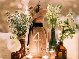 a lovely rustic wedding centerpiece of a wood slice, candles, white blooms and greenery in dark apothecary bottles and in a burlap sack