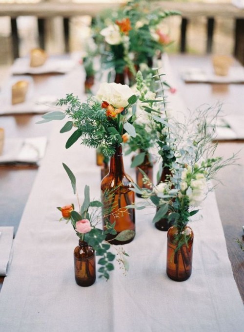 a white fabric table runner with dark apothecary bottles, with greenery and pink and white blooms is a lovely idea to rock for wedding table decor