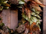 a pretty boho fall wedding wreath of lots of leaves and magnolia foliage, pinecones, wheat, bunny tails and feathers is a lovely and textural decor idea