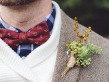 a stylish layered groom’s look with a blue plaid shit, a red printed bow tie, a white sweater, a brown blazer and a bold boutonniere