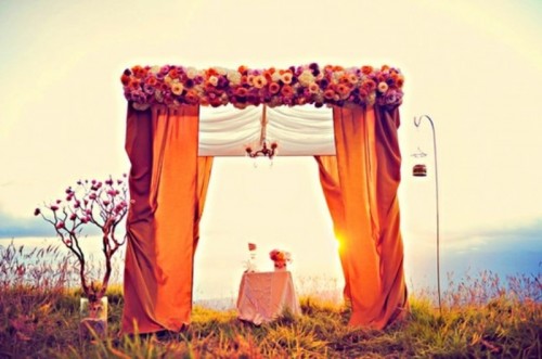 a super colorful wedding altar with burgundy and orange curtains, with bright blooms on top and a crystal chandelier