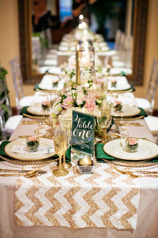 a stylish wedding tablescape with a white and gold glitter chevron table runner, gold glitter glasses and cutlery, neutral blooms, gold candles and green napkins