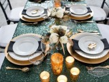 a shiny and bright wedding tablescape with a green sequin tablecloth, gold placemats, white porcelain, black napkins, candles and gold cutlery and neutral blooms