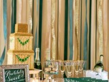 a shiny and chic wedding dessert and drink station, with a gold and emerald square wedding cake, gold-rimmed glasses, green cards, a green sign and a champagne bottle