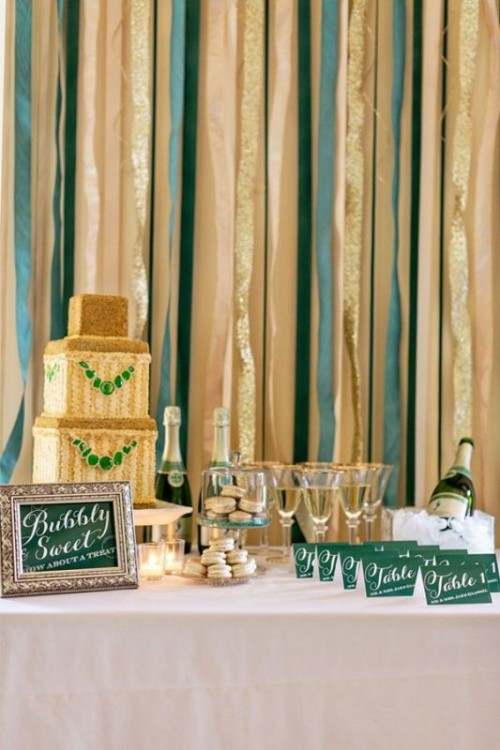a shiny and chic wedding dessert and drink station, with a gold and emerald square wedding cake, gold-rimmed glasses, green cards, a green sign and a champagne bottle