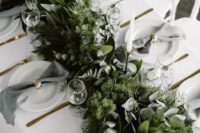 a chic neutral winter wedding tablescape with a super lush greenery runner with much texture, white porcelain, grey napkins and gold napkin rings