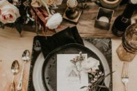 a moody winter wedding tablescape with elegant cutlery, black plates and placemats, gold candleholders and blush and burgundy blooms