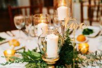 a refined winter wedding centerpiece of fresh greenery, pillar candles in candleholders, candles and a gold table number