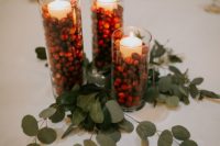 a simple winter wedding centerpiece of eucalyptus, cranberries and pillar candles in tall glasses looks bold and chic
