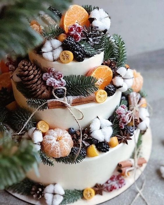 a white wedding cake topped with a whole number of stuff - citrus, berries, pinecones, cinnamon bark, cotton and evergreens
