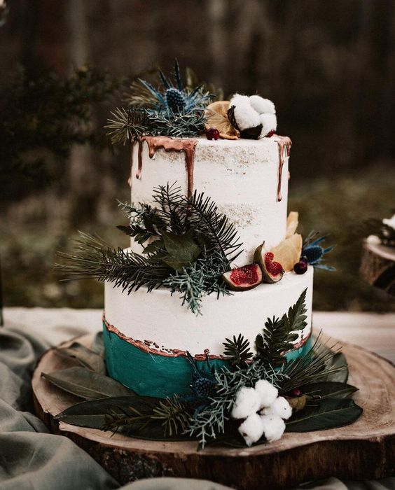 a white wedding cake with an emerald detail, evergreens and foliage, cotton, thistles and some fruit slices is amazing for winter
