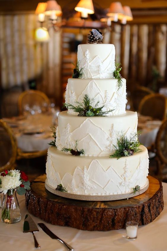 a white wedding cake with painted mountains and trees, with greenery and berries and lovely pinecone toppers