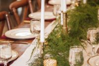 a winter holiday wedding table with pine, gilded plates and candleholders, burgundy napkins and thin and tall candles