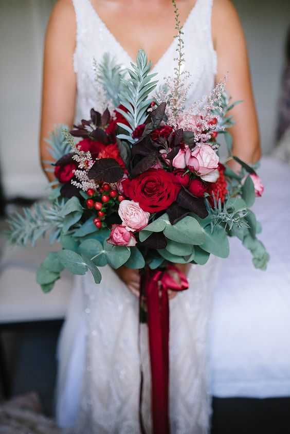 a bold and dimensional wedding bouquet with dark leaves, red and pink blooms, eucalyptus and other greenery and red ribbons