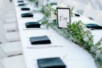 a fresh wedding tablescape with a white tablecloth, black napkins, a lush greenery and white bloom runner and a table number