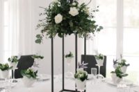 a stylish minimalist wedding tablescape with a tall centerpiece on a black stand, lush white blooms and greenery and black napkins