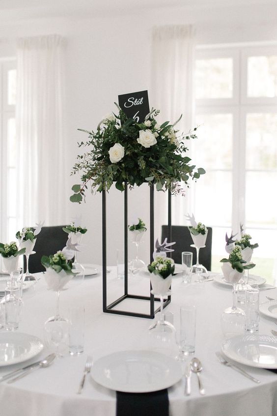 a stylish minimalist wedding tablescape with a tall centerpiece on a black stand, lush white blooms and greenery and black napkins