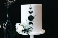 a white wedding cake decorated with black moon phases and white blooms and greenery is ideal for a celestial wedding