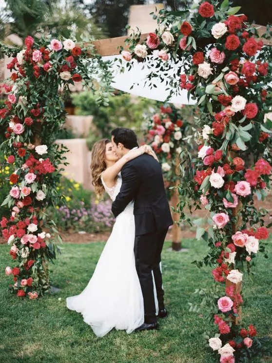 a bold wedding arch of red, blush and white blooms and greenery and sheer fabric is a chic idea