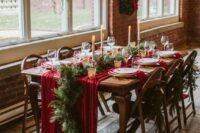 a lovely winter wedding reception space with evergreens and lights, a red table runner and evergreens and tall candles is chic