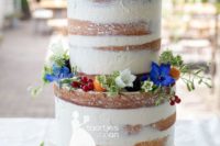 a naked wedding cake with colorful and white blooms, berries and fruits and some Lego toppers