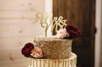 a refined and glam wedding cake with a gold glitter tier, a white one and a white flower one, with gold drip, burgundy and pink blooms and a calligraphy topper