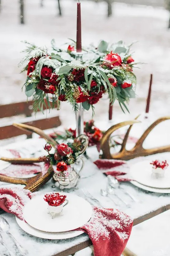a rustic winter wedding tablescape with greenery and red and burgundy blooms, antlers, red linens for a woodland feel
