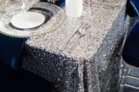 a silver glitter wedding tablecloth paired with navy anpkins, white blooms and candles and silver cutlery for a super glam and shiny tablescape