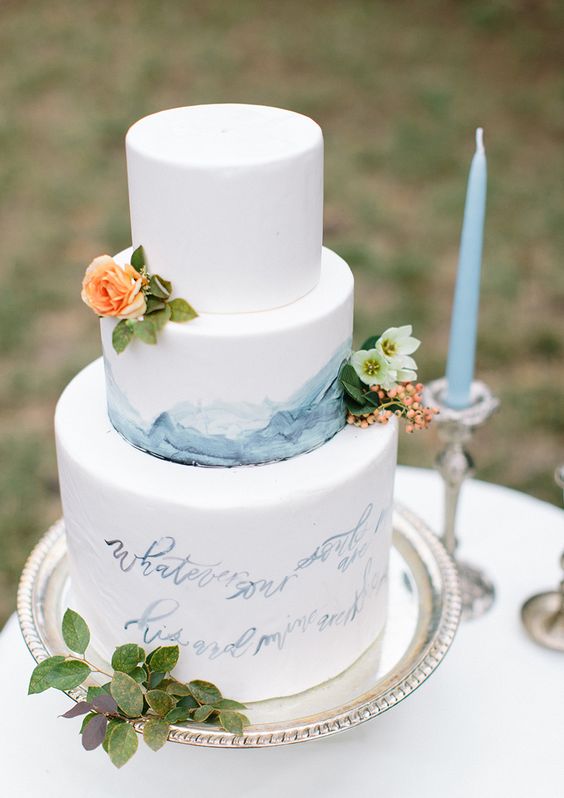 a white buttercream wedding cake with blue watercolors and blue love letters, neutral and bright blooms and greenery