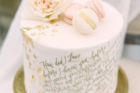 a white one-tier wedding cake with gold leaf and a gold calligraphy love letter, with peachy and white macarons and a peachy bloom