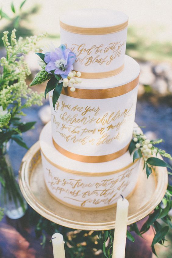 a white wedding cake with copper ribbon and gold calligraphy love letters on the tiers, a blue bloom and some flowers is a lovely idea for a spring wedding