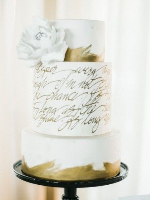 a white wedding cake with gold leaf tiers and a love letter one in the center, a white sugar bloom is a lovely idea for a chic and cool wedding