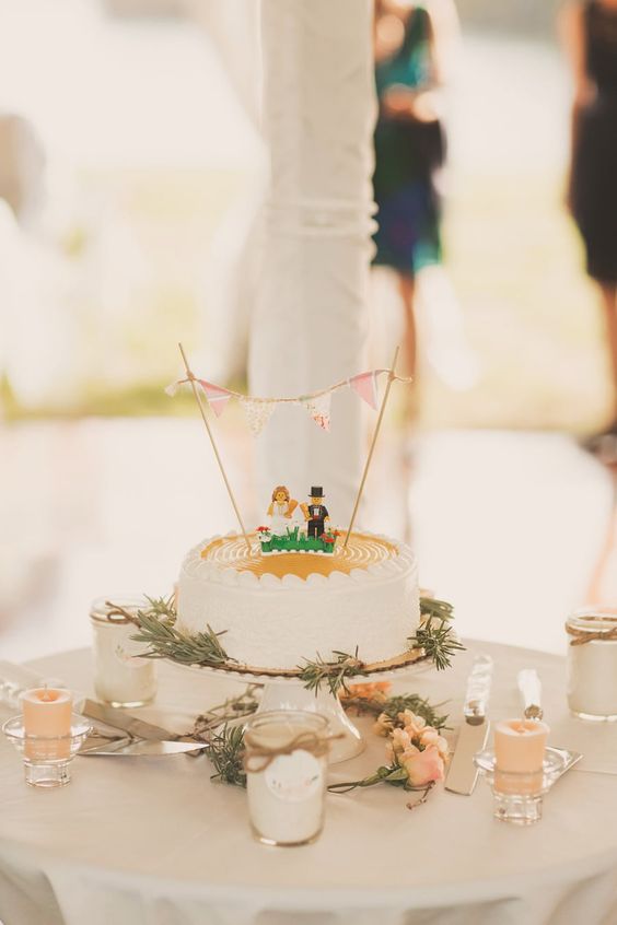 a white wedding cake with greenery, Lego figurines and a banner on top for a fun and cool look