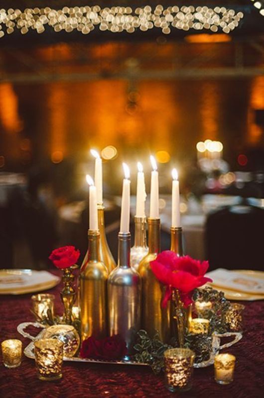 a chic and refined wedding centerpiece of gilded bottles, tall and thin candles, red roses and mercury glass candleholders and some foliage