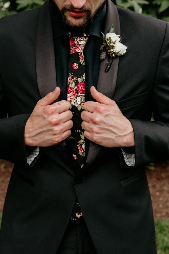 a black tuxedo with shiny lapels, a black shirt and a moody floral tie plus a white floral boutonniere are a lovely combo for a wedding