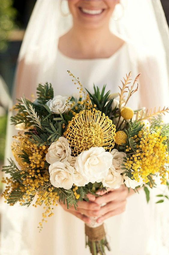 a bold yellow wedding bouquet of pincushion proteas, billy balls, mimosas, white roses and lots of greenery is wow