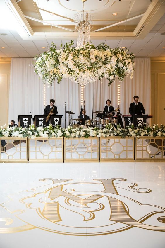 a chic and refined gold and white dance floor with monograms, with an oversized chandelier with greenery and white blooms over it