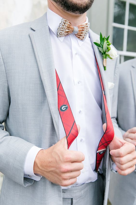 a grey suit, a white shirt, a printed bow tie, red striped suspenders and a greenery boutonnire