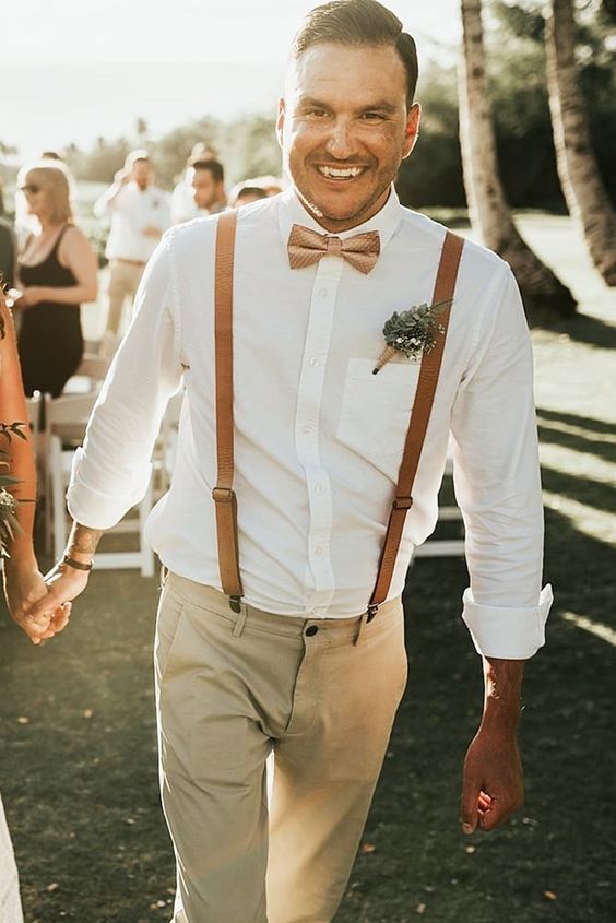 a light summer groom's look with a white shirt, amber leather suspenders, tan pants and a greenery boutonniere