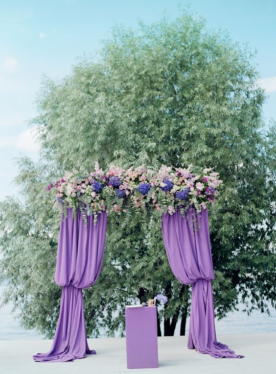 a purple wedding arch with curtains, lush blooms and greenery looks very eye-catching