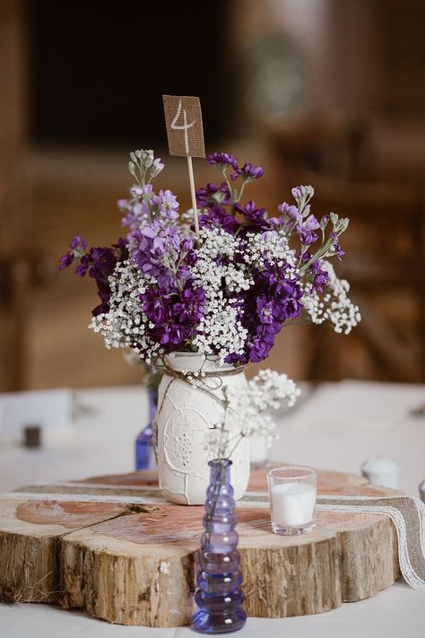 a white mason jar centrepiece with purple and white flowers and a burlap table number