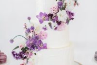 a white wedding cake topped with purple and blush blooms plus some leaves