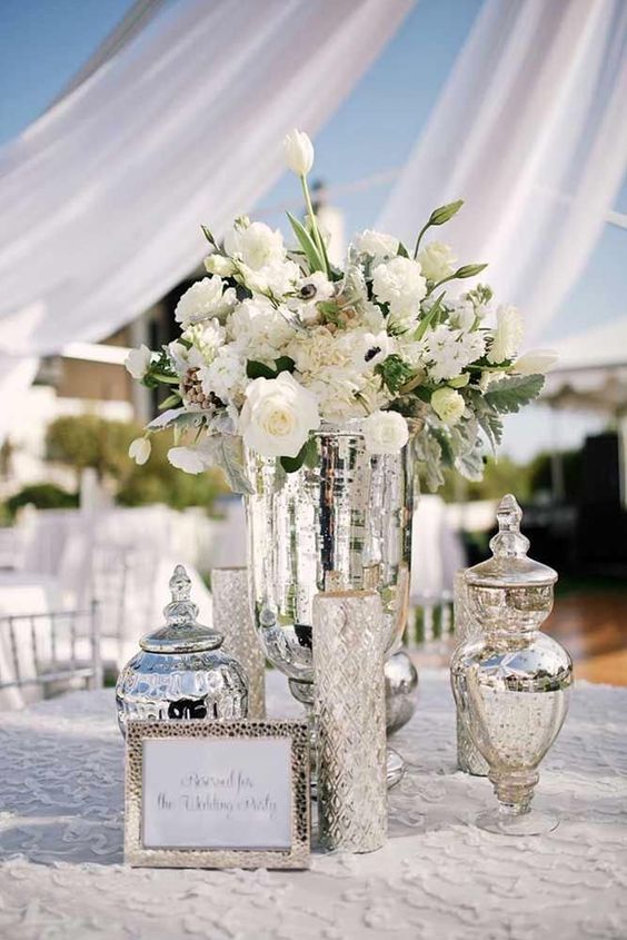 An elegant and shiny silver wedding centerpiece of mercury glass jars, vases and white lush blooms