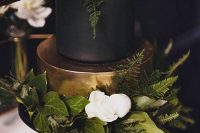 a refined and chic matte black and shiny gold wedding cake with lush greenery and white roses is elegant