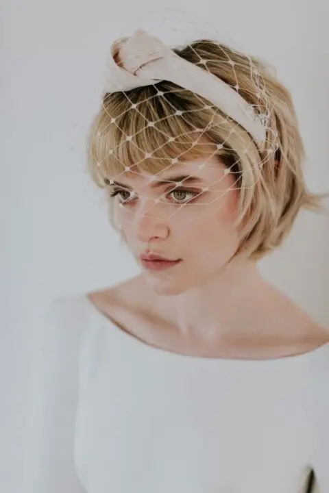 a short blonde bob with bangs, a knot headband and a birdcage veil for a retro touch to the look