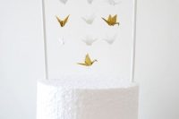 a white buttercream wedding cake with a stand and lots of small white and gold paper cranes hanging down from it is a very cute idea