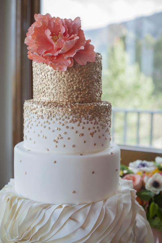 a white wedding cake covered with gold polka dots and a coral sugar bloom on top