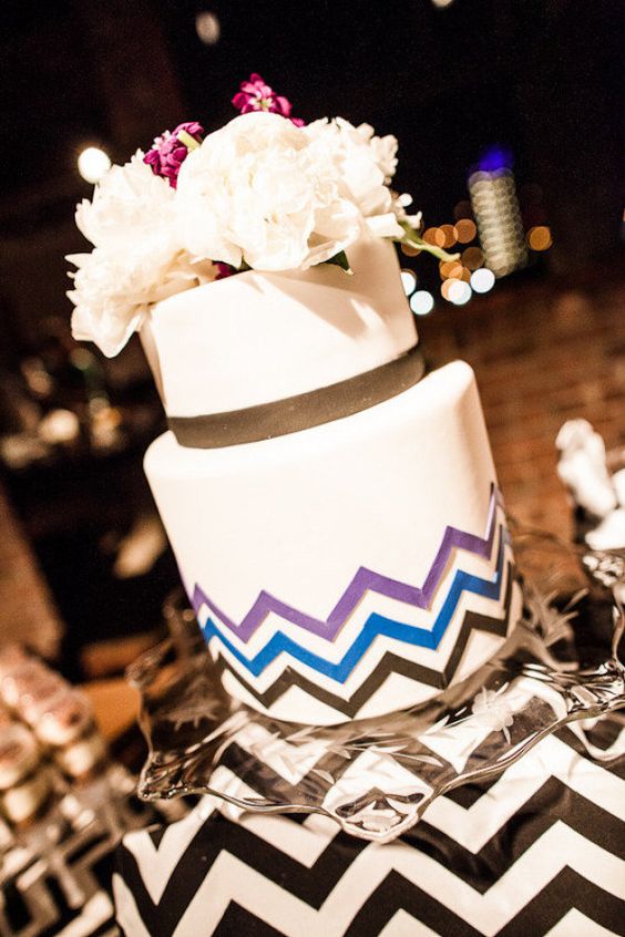 a white wedding cake with black ribbon, purple, blue and black chevron decor and fresh blooms on top is a lovely idea for a mid-century modern wedding