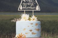 an adventure-themed wedding cake with a gold tier and a white one with gold leaf, fresh blooms, grass and a plywood cake topper